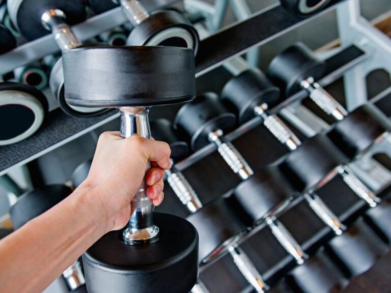 Hand hält eine Kurzhantel vor einem Hantelständer – Kurzhanteltraining für Kraftaufbau und Ganzkörperübungen.